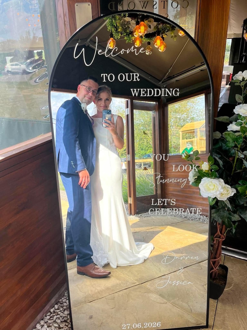 May include: An arched floor mirror with a black frame featuring white text that reads Welcome TO OUR WEDDING YOU LOOK Stunning LET'S CELEBRATE Darren & Jessica 27.06.2026. The mirror reflects a person in a blue suit and a person in a white dress.