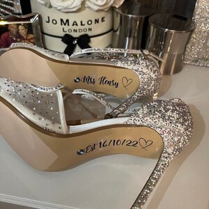 May include: A pair of white sparkly high heel shoes with the text "Mrs. Henry" and "Est 16/10/22" written on the soles.