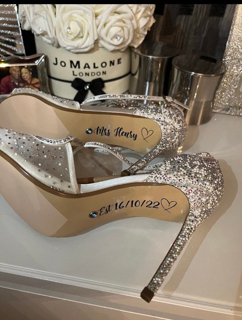 May include: A pair of white sparkly high heel shoes with the text "Mrs. Henry" and "Est 16/10/22" written on the soles.