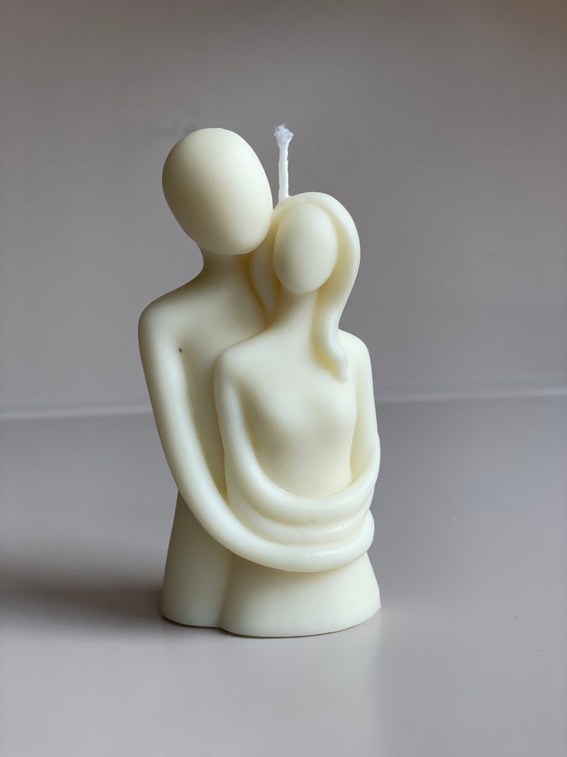 May include: White candle sculpture of two figures embracing. The figures are stylised and abstract, with smooth, rounded forms. The candle is a romantic and modern home decor piece.