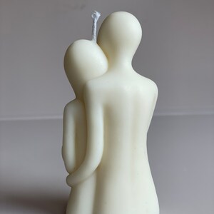May include: A white candle sculpted in the shape of two figures embracing. The candle has a single wick.