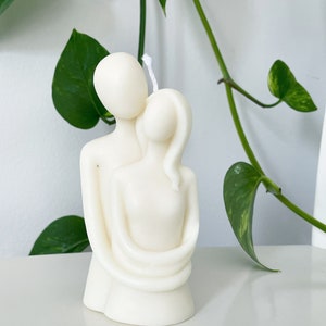 May include: White candle sculpture of two figures embracing. The figures are stylised and abstract, with simple, rounded forms. The candle is made of a smooth, white wax.