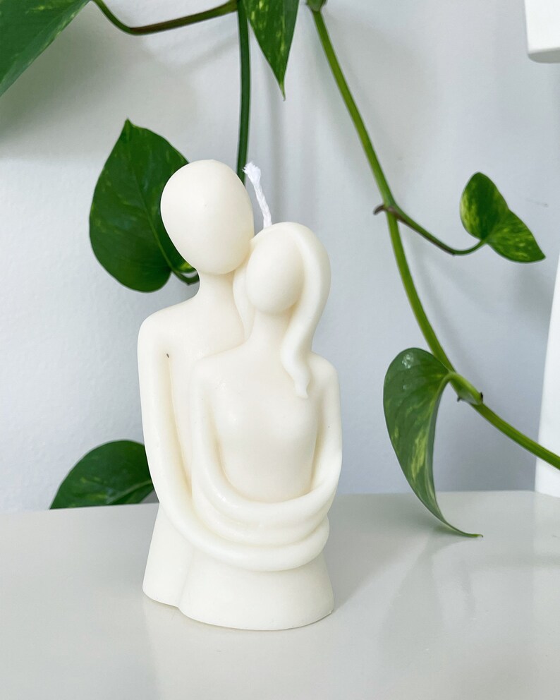 May include: White candle sculpture of two figures embracing. The figures are stylised and abstract, with simple, rounded forms. The candle is made of a smooth, white wax.