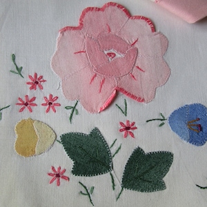 May include: A close-up of a white fabric with embroidered flowers. The flowers are pink, blue, yellow, and green. The fabric has a delicate texture.