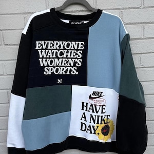 May include: Color-blocked Nike sweatshirt with the text "EVERYONE WATCHES WOMEN'S SPORTS" and "HAVE A NIKE DAY." The design includes black, white, blue, and green panels. A sunflower graphic is also present.