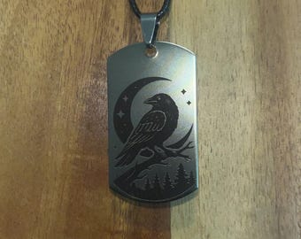 May include: A silver-colored dog tag pendant with a black cord. The tag features an engraved design of a raven perched on a branch with a crescent moon and stars. The pendant is rectangular.