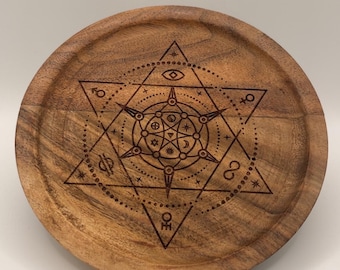 May include: A round wooden tray with a detailed engraved geometric design. The design features a central star with surrounding symbols and patterns. The tray is made of a light brown wood.