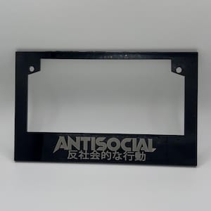 May include: Black license plate frame with the word "ANTISOCIAL" and Japanese text. The frame has a rectangular shape with rounded corners and two mounting holes.
