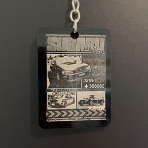 May include: Black rectangular keychain with a silver chain and ring. The keychain features a Subaru design with images of cars and Japanese text. The word "SUBARU" is at the top of the keychain.
