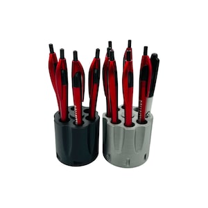 May include: Two pen holders, one black and one gray, each with six cylindrical openings. The holders contain red pens and a black marker. The pens have a sleek design. The text "BALLISTICOS" is printed on the pens.