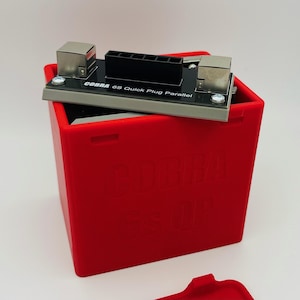 May include: A red, square container with the text "COBRA 6S Quick Plug Parallel" printed on a gray and black device on top. The container also has the text "COBRA 6S UP" printed on the side. The device has two silver metal blocks.