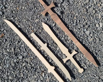 May include: Five wooden toy swords of varying sizes and designs are arranged on a gray, gravel surface. The swords are made of light-colored wood, with one sword in a darker wood tone. The swords have different handle and blade shapes.