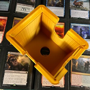 May include: A yellow, 3D-printed card holder, designed with an angled shape and a circular opening. The holder is positioned over a collection of trading cards, suggesting its use for organizing or displaying the cards. The holder's design is simple and functional.