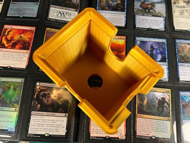 May include: A yellow, 3D-printed card holder, designed with an angled shape and a circular opening. The holder is positioned over a collection of trading cards, suggesting its use for organizing or displaying the cards. The holder's design is simple and functional.