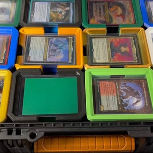 May include: A black and orange storage case with multiple compartments, each holding trading cards and dice. The compartments are various colors, including blue, yellow, green, and white, with clear plastic lids. The case is designed for organizing and protecting collectible cards.