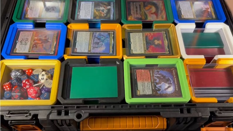 May include: A black and orange storage case with multiple compartments, each holding trading cards and dice. The compartments are various colors, including blue, yellow, green, and white, with clear plastic lids. The case is designed for organizing and protecting collectible cards.