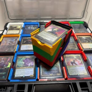 May include: A black storage case with multiple compartments, each holding trading cards in protective sleeves. A multi-colored card holder sits atop the case, filled with more cards. The case has a clear lid.