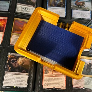 May include: A gold-colored, 3D-printed card holder, open to reveal a stack of blue-backed cards. The holder is rectangular with a lid and is surrounded by various trading cards in a binder.