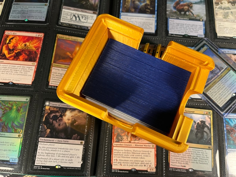 May include: A gold-colored, 3D-printed card holder, open to reveal a stack of blue-backed cards. The holder is rectangular with a lid and is surrounded by various trading cards in a binder.