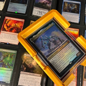 May include: A gold-colored card holder with a clear plastic cover, holding a Magic: The Gathering card. The card features the text "Shorikai, Genesis Engine" and has a blue and white illustration. The holder is designed to protect the card.