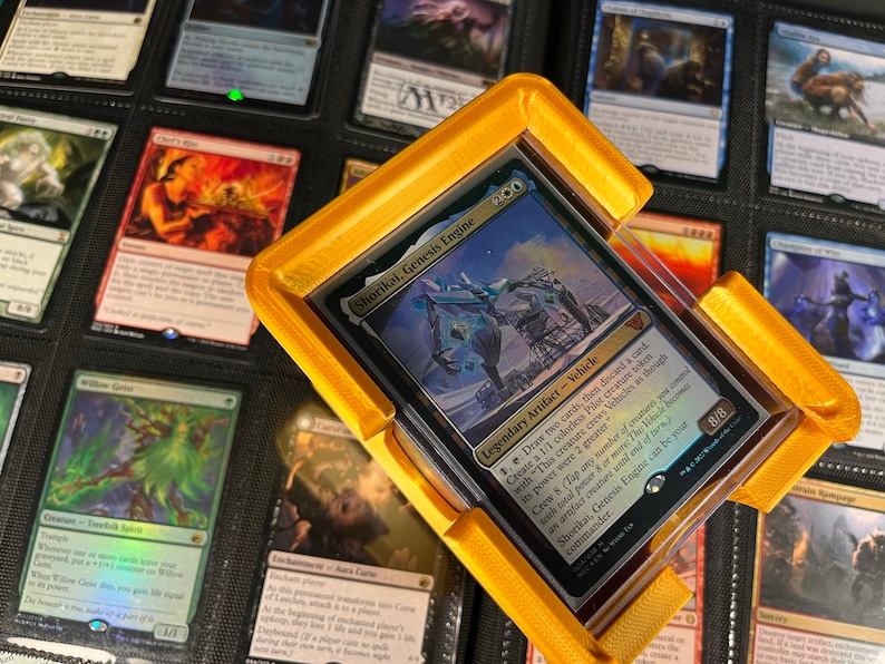 May include: A gold-colored card holder with a clear plastic cover, holding a Magic: The Gathering card. The card features the text "Shorikai, Genesis Engine" and has a blue and white illustration. The holder is designed to protect the card.