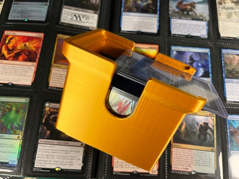 May include: A gold-colored, 3D-printed card holder with a lid, designed to hold trading cards. The holder is open at the top and has a curved design. The holder is sitting in front of a binder filled with trading cards.