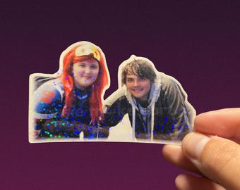May include: A clear sticker featuring two people, one with red hair and the other with brown hair, both smiling. The sticker has a glittery effect with blue and silver specks. The text "cassie makes art." is at the bottom.