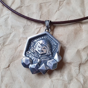 May include: A silver pendant necklace with a detailed, stylized figure with a mask design. The pendant is attached to a brown leather cord. The pendant has a geometric shape with several geometric shapes attached to the bottom.