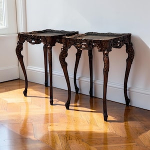 May include: A pair of dark brown wooden side tables featuring square tops with inset surfaces and intricate carved detailing along the aprons. Each table stands on four slender, curved cabriole legs that terminate in detailed feet.