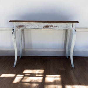 May include: A distressed white and brown console table with curved legs. The table has a dark brown top and a white-painted body with visible wear and tear, giving it a vintage appearance. The table is set against a white wall and a wooden floor.