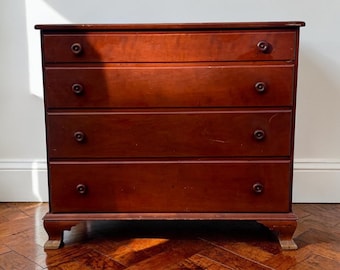 May include: A four-drawer wooden dresser with a rich, reddish-brown finish. The dresser has round, dark knobs and sits on short, curved feet. The wood grain is visible, and the piece appears to be well-maintained.