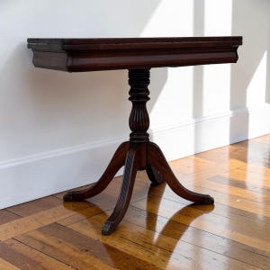 May include: A dark brown wooden pedestal table with a rectangular top and a central column base. The table features a fluted pedestal support that transitions into four curved legs, each ending in a small, rounded foot detail.
