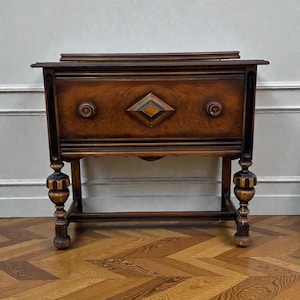 May include: A dark brown antique wooden cabinet with a diamond-shaped inlay and round knobs. The cabinet has ornate legs and a lower shelf. The wood grain is visible throughout the piece, and it sits on a parquet floor.