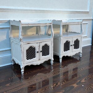 May include: Two matching white distressed bedside tables. Each table has a shelf, a cabinet with a wire mesh door, and decorative legs. The tables are painted white with distressed edges, giving them a vintage look.