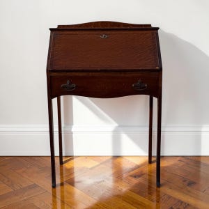 May include: A dark brown wooden slant-front desk featuring a hinged lid with a central metal keyhole escutcheon. The desk includes a single drawer positioned below the writing surface, accented with two decorative metal pull handles.