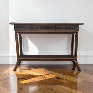 May include: A brown rattan console table featuring a rectangular top, a central drawer with a small knob, and a lower shelf. The frame is constructed from curved rattan poles, while the drawer front and lower shelf display a woven pattern.