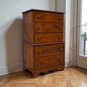 May include: A two-piece wooden dresser with a warm brown finish. The top section has four drawers, and the bottom section has two drawers. Each drawer has a round wooden knob. The dresser is set against a neutral wall and sits on a parquet floor.