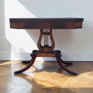 May include: A dark wood game table featuring a rectangular top with rounded corners and a central pedestal base shaped like a lyre with three vertical metal rods. The base is supported by four curved legs with dark feet and decorative inlay details.