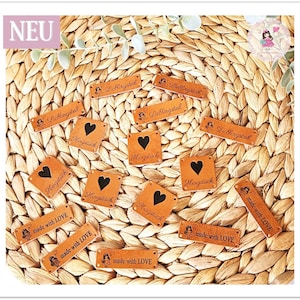 Può includere: Una collezione di etichette in pelle marrone con vari design. Alcune etichette presentano il testo "Lieblingsteil" e altre presentano il testo "Herzstück" con un design a forma di cuore. Alcune etichette presentano anche il testo "made with LOVE".