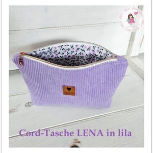 Könnte beinhalten: Eine lavendelfarbene Kosmetiktasche aus Cord mit floralem Innenfutter. Sie hat einen goldenen Reißverschluss und ein kleines braunes Lederetikett mit einem Herz. Der Text "Cord-Tasche LENA in lila" befindet sich unten.