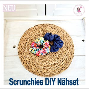 Könnte beinhalten: Zwei Stoff-Haargummis, eines mit blau-weißem Punkte-Muster und eines mit buntem Blumenmuster, auf einer geflochtenen Strohhutmatte. Der Text "Scrunchies DIY Nähset" ist unter der Matte gedruckt.