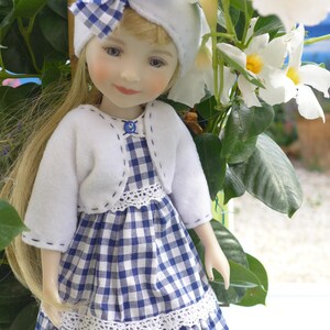 Peut inclure: Poupée en porcelaine aux longs cheveux blonds, portant un bandeau blanc avec un nœud vichy bleu et blanc. La poupée porte une veste boléro blanche sur une robe vichy bleue et blanche avec une bordure en dentelle blanche. La poupée est posée près de fleurs blanches.
