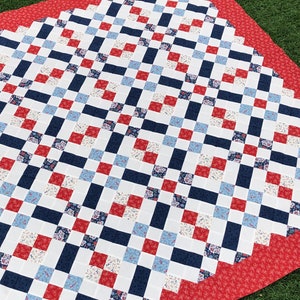 Puede incluir: Un edredón hecho a mano con un patrón repetido de cuadrados rojos, blancos y azul marino. Algunos cuadrados presentan diseños florales. El edredón tiene un borde rojo con un pequeño diseño floral blanco. El edredón se muestra sobre una superficie verde.