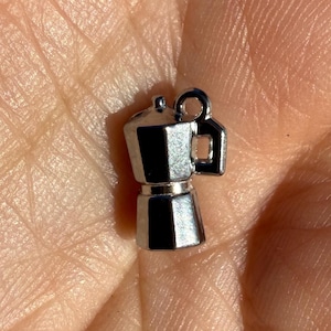 May include: A miniature silver-toned metal charm shaped like a traditional stovetop espresso maker. The charm features a classic octagonal base, a central waist, and a small handle on the side, topped with a small circular loop for attachment.
