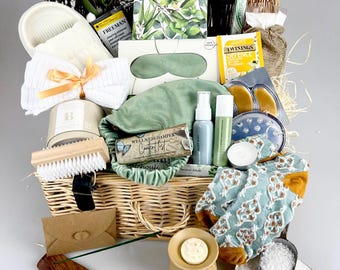 May include: A wicker basket filled with self-care items. Includes a sleep mask, bath salts, candles, tea, socks, a brush, and a wooden incense holder. The basket is decorated with greenery and pinecones, creating a spa-like gift set.