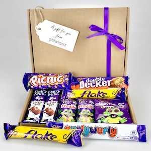 Puede incluir: Una caja de regalo de cartón marrón con una cinta morada y una etiqueta de regalo blanca que dice "Un regalo para ti de gifthamperz". La caja está llena de diferentes barras de chocolate, incluyendo Cadbury Dairy Milk, Cadbury Flake, Cadbury Picnic, Cadbury Double Decker, Cadbury Freddo y Cadbury Crunchie.