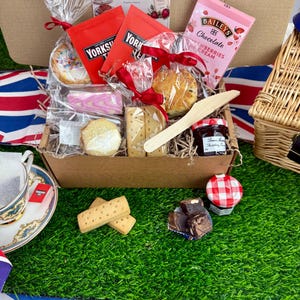 May include: A cardboard gift box containing Yorkshire Tea bags, a Baileys Strawberries and Cream chocolate bar, a scone, shortbread, a small jar of Bonne Maman raspberry preserves, and a wooden knife. A card reads Wimbledon at Home Afternoon Tea.