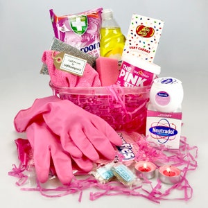 May include: A pink and white gift basket filled with cleaning supplies, sweets, and pink rubber gloves. The basket includes a bottle of washing up liquid, a pink cleaning cloth, a pink scouring pad, a pack of cleaning wipes, a pink star-shaped cleaning product, a pack of sweets, a pink scented candle, and a pink rubber glove.