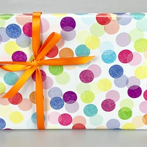 May include: A rectangular gift box wrapped in white paper with a colorful polka dot pattern in shades of red, purple, blue, yellow, and orange. The box is tied with a bright orange satin ribbon and bow.