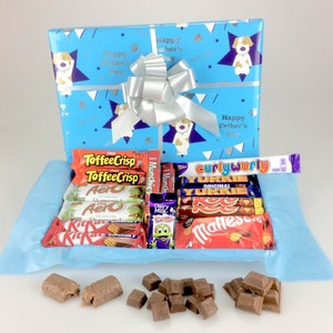 May include: A gift box wrapped in blue paper with a silver bow, featuring a variety of chocolate bars. The wrapping paper has a pattern of stars, paw prints, and the text "Happy Father's Day". Assorted chocolate bars are visible inside the box.